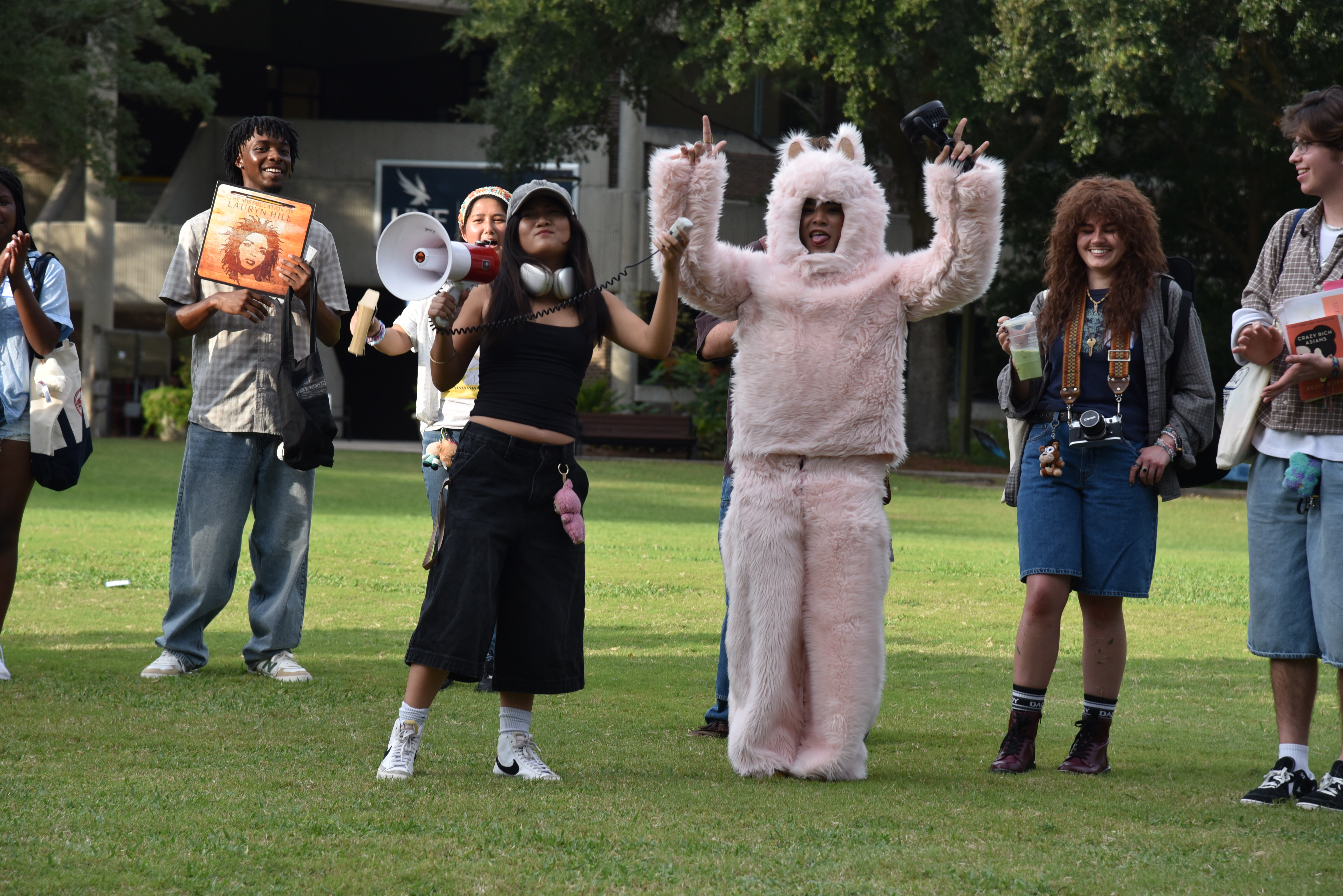 A student announces a contestant dressed in a full-size Labubu costume as the winner. The contestant stands with their arms raised in the air.